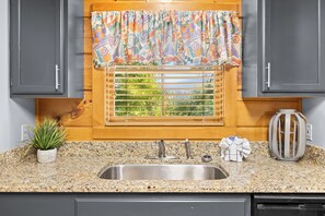 A kitchen with a granite countertop, stainless steel sink, wooden cabinets, a window with a patterned curtain, and a potted plant.
