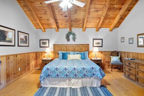 Une chambre avec un plafond en bois, un lit avec une literie à motifs, deux tables de chevet avec des lampes, une commode et des tableaux encadrés accrochés aux murs.