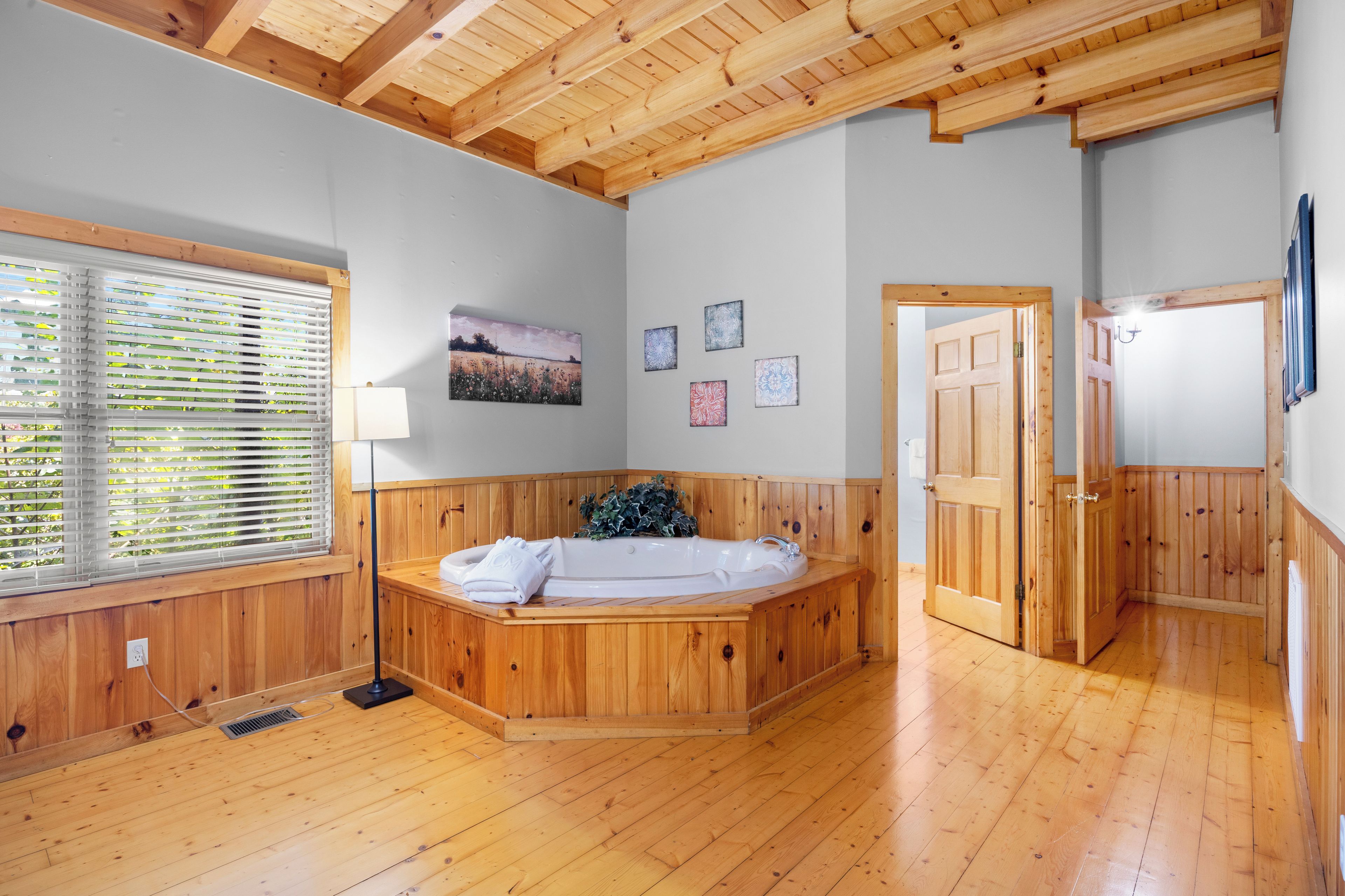 Une pièce avec un plancher en bois, une grande fenêtre avec des stores, une baignoire en bois et une porte donnant sur une autre pièce.