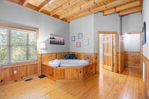Une pièce avec un plancher en bois, une grande fenêtre avec des stores, une baignoire en bois et une porte donnant sur une autre pièce.