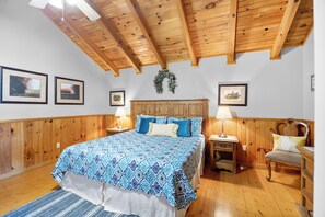 Une chambre avec un plafond en bois, un lit recouvert d’une literie à motifs, deux tables de chevet avec des lampes et des tableaux encadrés accrochés aux murs.