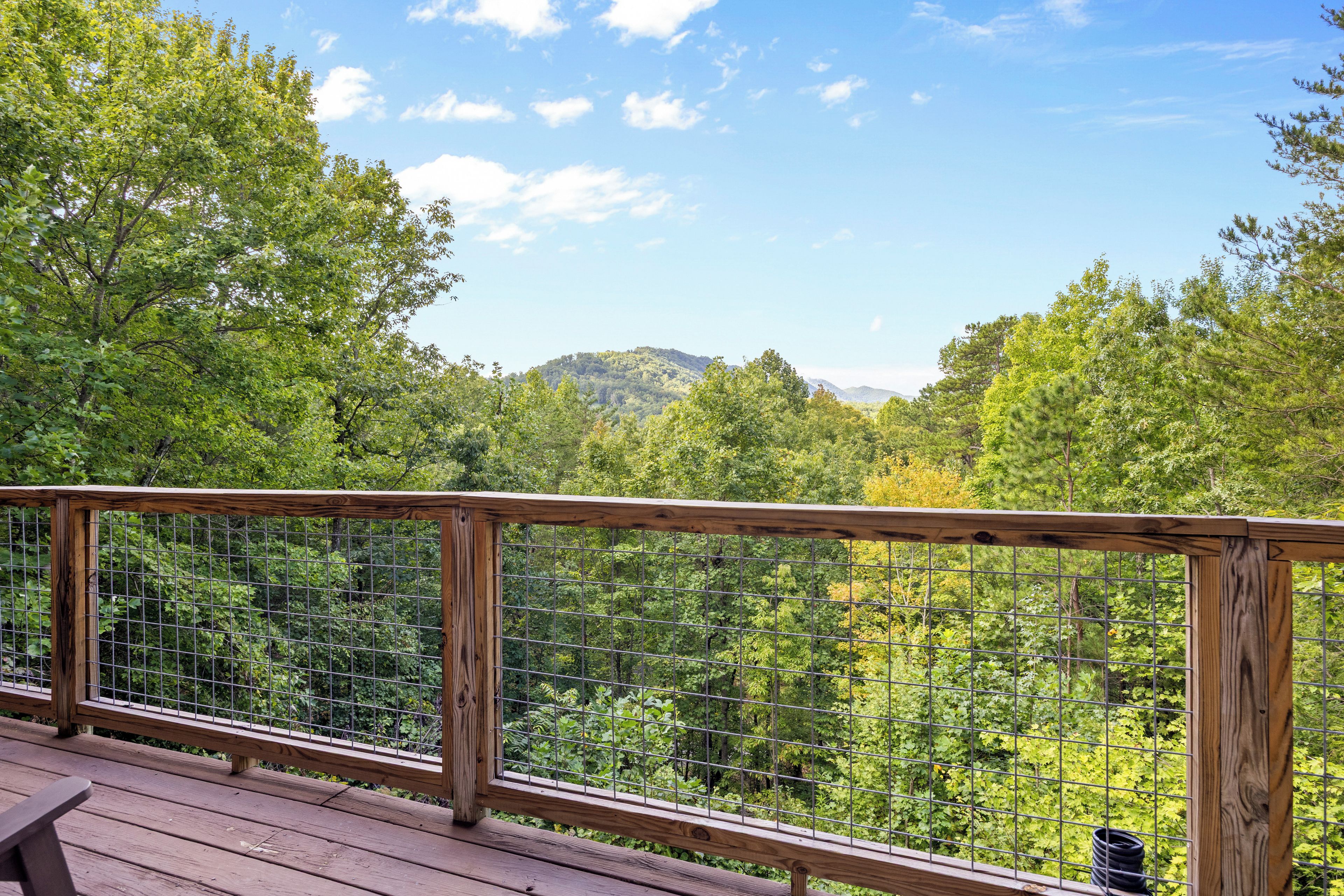 Une terrasse en bois, dotée d’une rambarde en métal, offre une vue sur une zone boisée avec des collines à l’horizon.