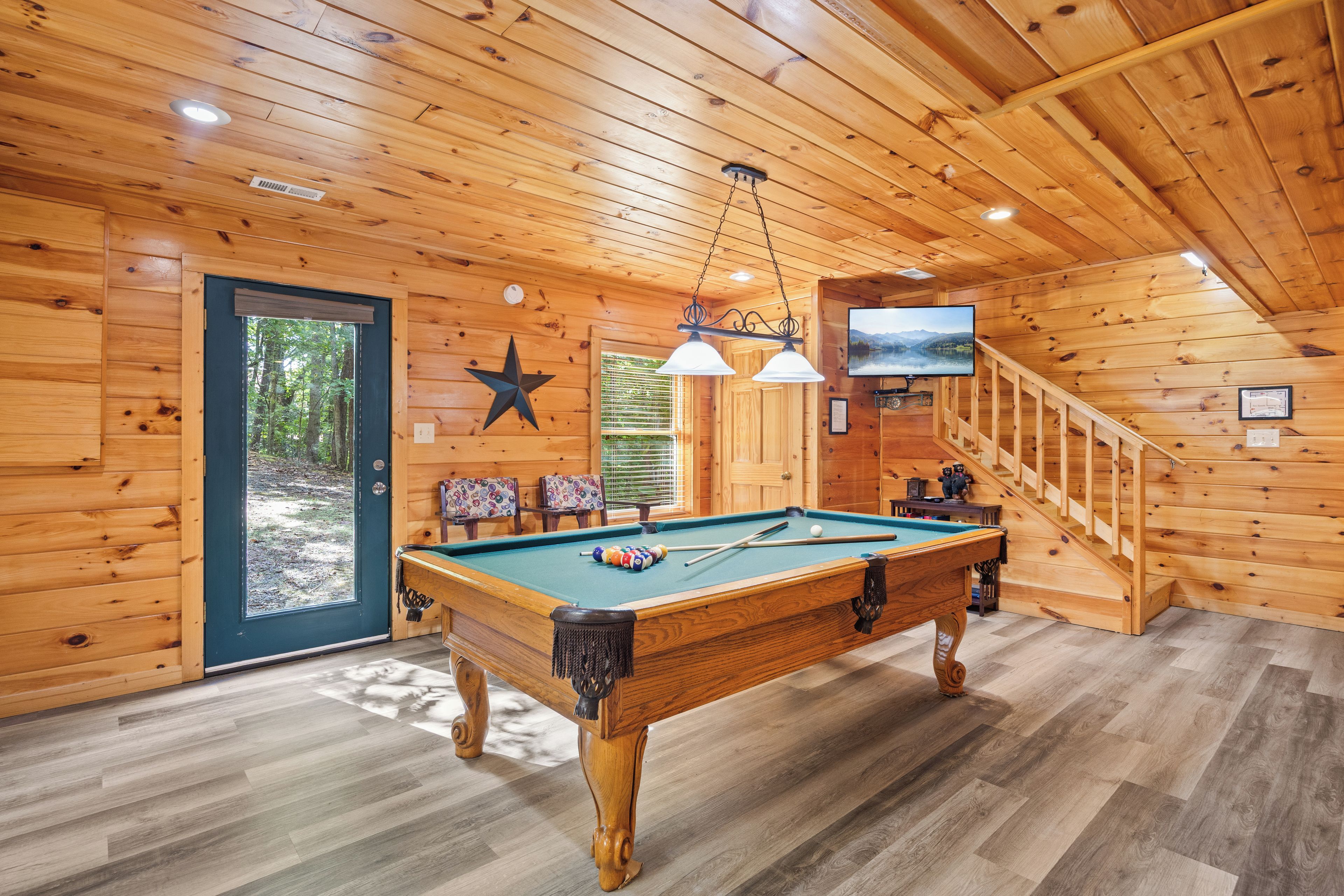 A wooden interior with a pool table, a television mounted on the wall, and a staircase.