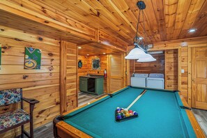 Une table de billard avec des billes, un canapé vert et un plafond en bois avec un luminaire suspendu.