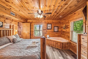 A wooden cabin bedroom with a bed, a window, a bathtub, and a ceiling fan. - Three Bears-  Enjoy a panoramic view of the Cove Mountain range! 2 bed/2 bath cabin! (Gatlinburg)