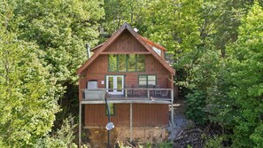 A two-story wooden cabin with a balcony, surrounded by dense forest. - Three Bears-  Enjoy a panoramic view of the Cove Mountain range! 2 bed/2 bath cabin! (Gatlinburg)