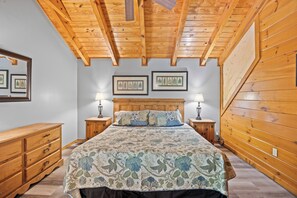 Une chambre avec un plafond en bois, un lit recouvert d’une couvre-lit à motifs floraux, deux tables de chevet avec des lampes et une commode en bois.