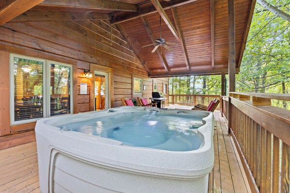 A hot tub area with a wooden deck, outdoor seating, and a view of the surrounding trees.