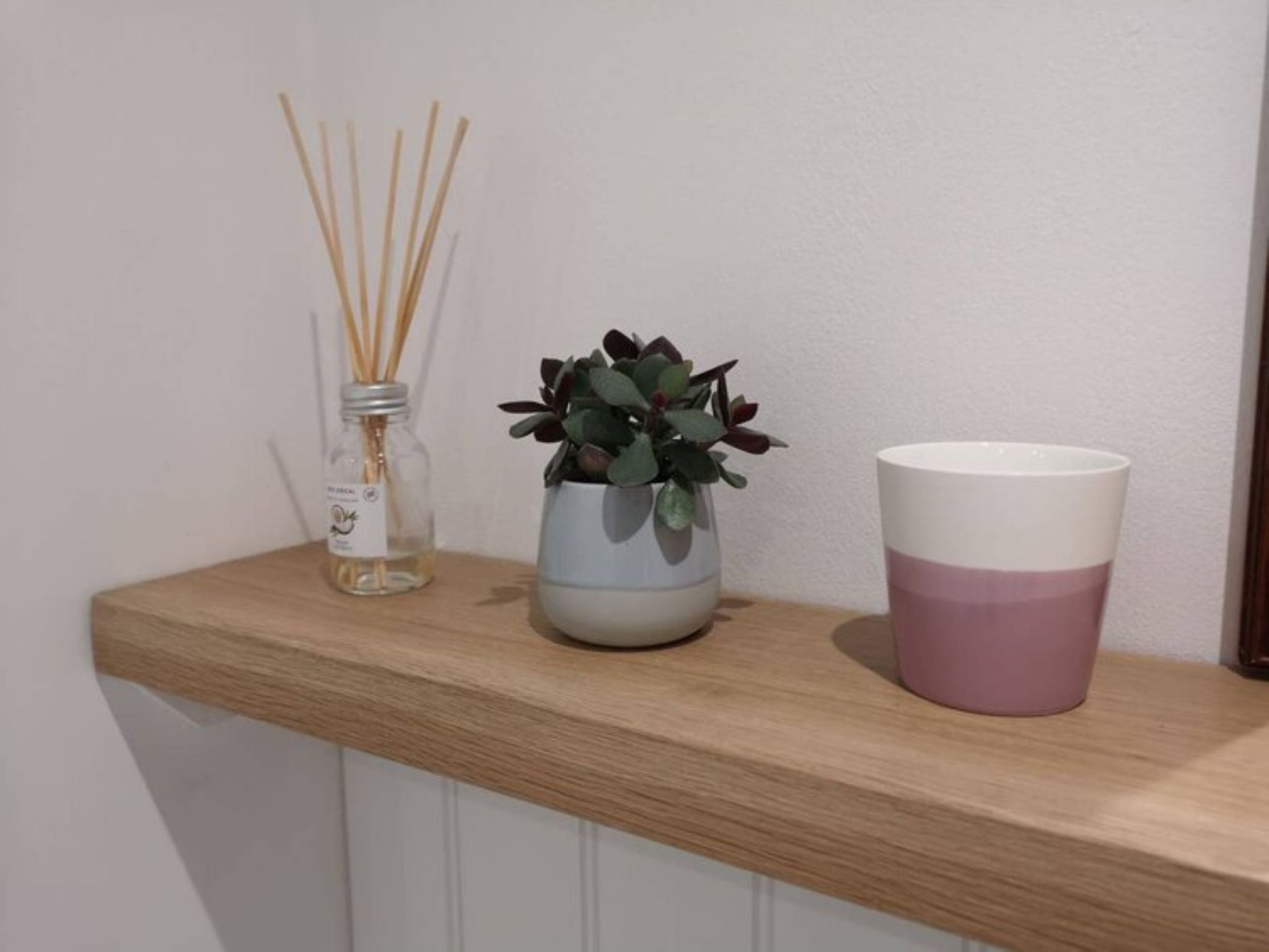 A wooden shelf with a glass jar of reed sticks, a potted plant, and a two-tone cup.