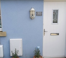 Uma casa com fachada azul, porta branca, placa com o número 80, um lampião e um pequeno vaso de planta.