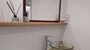 A modern bathroom with a glass bowl sink, a wooden vanity, a mirror above the sink, and a shelf with a plant and reed diffuser. - The Little House, Boutique Home Near the sea (Chichester)