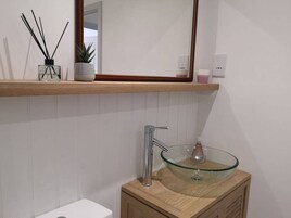Uma casa de banho moderna com lavatório em vidro, toucador de madeira, espelho sobre o lavatório e uma prateleira com uma planta e difusor de aroma a caniço.