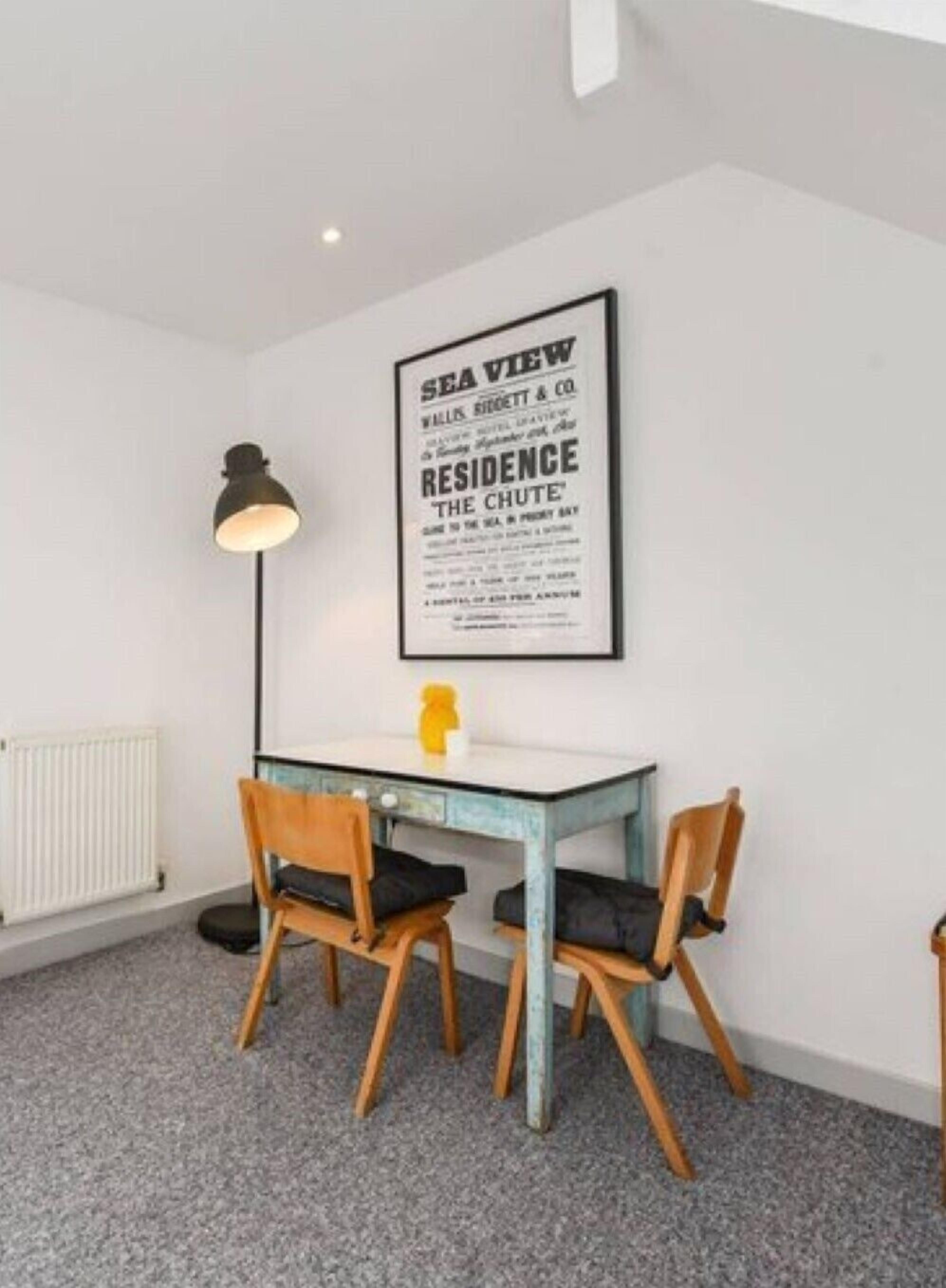 A room with a white wall, a framed poster, a table, two chairs, a floor lamp, and a radiator.