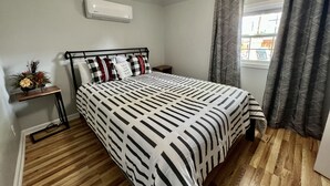 Ein Schlafzimmer mit einem Bett, einem Nachttisch, einer Wand-Klimaanlage, einem Fenster mit Vorhängen und einer kleinen Ablage mit einer Pflanze.
