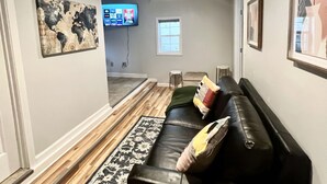 Ein Wohnzimmer mit einem schwarzen Ledersofa, einem gemusterten Teppich, einem flachbildfernseher an der Wand und einem gerahmten Bild über dem Sofa.