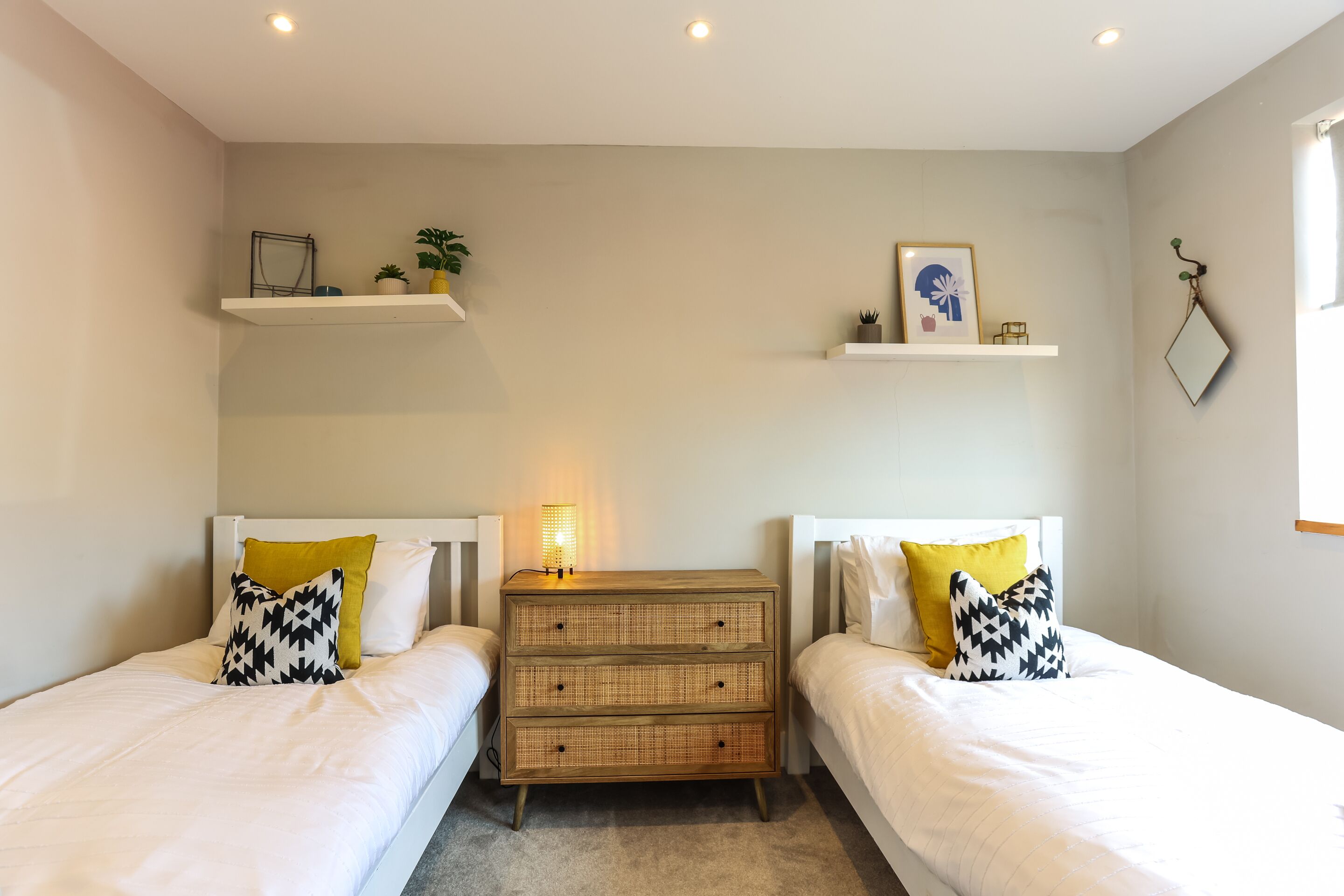 A bedroom with two single beds, a bedside table with a lamp, and shelves with decorative items.