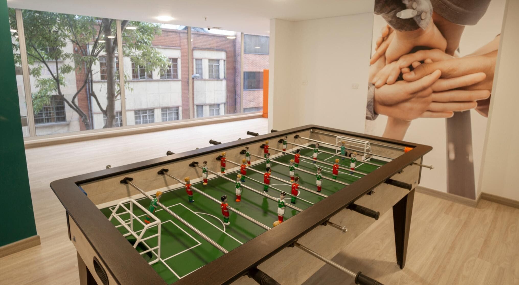 A foosball table in a room with a large window, a mural of hands, and a view of buildings outside.