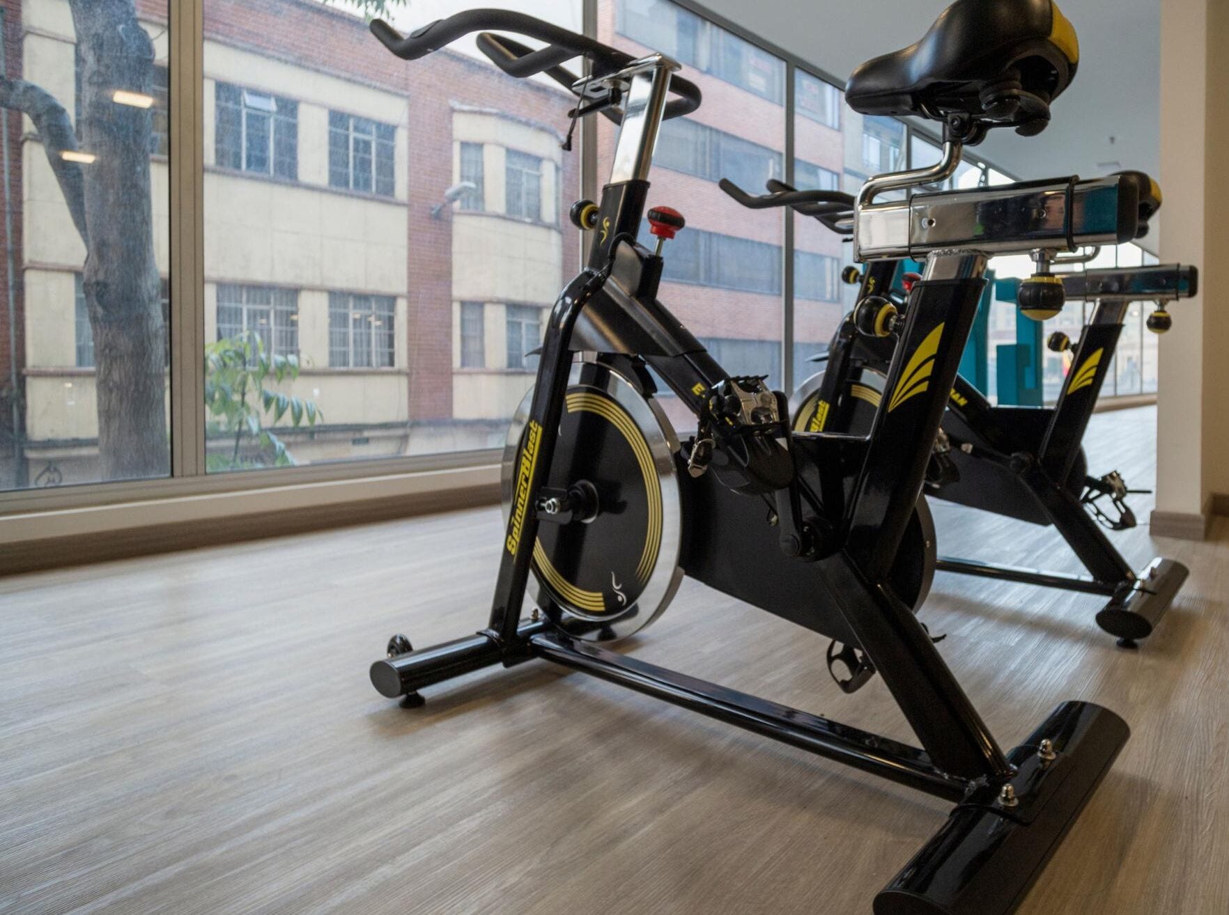 Een stilstaande fiets in een kamer met grote ramen, een houten vloer en uitzicht op een gebouw.