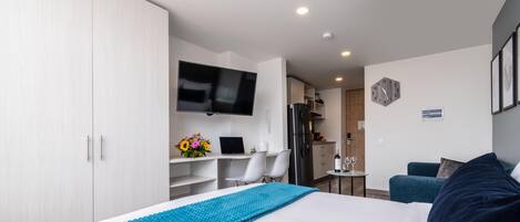 A modern hotel room with a large bed, a desk with a laptop, a TV, and a kitchenette in the background.