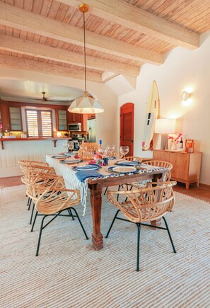 A dining area with a wooden table set for six, surrounded by wicker chairs, a hanging light fixture, and a surfboard on the wall. - Villa Rosso Di Sera: 2 Minute Walk from the Beach (Chalk Sound)