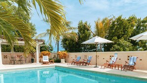 Ein Swimmingpool mit Liegestühlen und Sonnenschirmen, umgeben von Palmen und blauem Himmel.