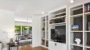 Una sala de estar moderna con televisor, estanterías blancas y un comedor con sillas y una mesa.