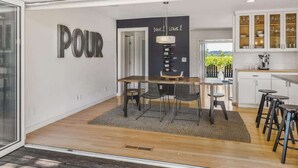 Una cocina moderna con armarios blancos, una zona de comedor con mesa y sillas de madera, y un cartel en la pared que dice 'POUR'.