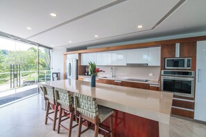 A modern kitchen with a central island, bar stools, stainless steel appliances, and a view of greenery outside. - Luxury Villa #10 Private Pool, Oceanview, & Complimentary Concierge Services (Playa Hermosa)