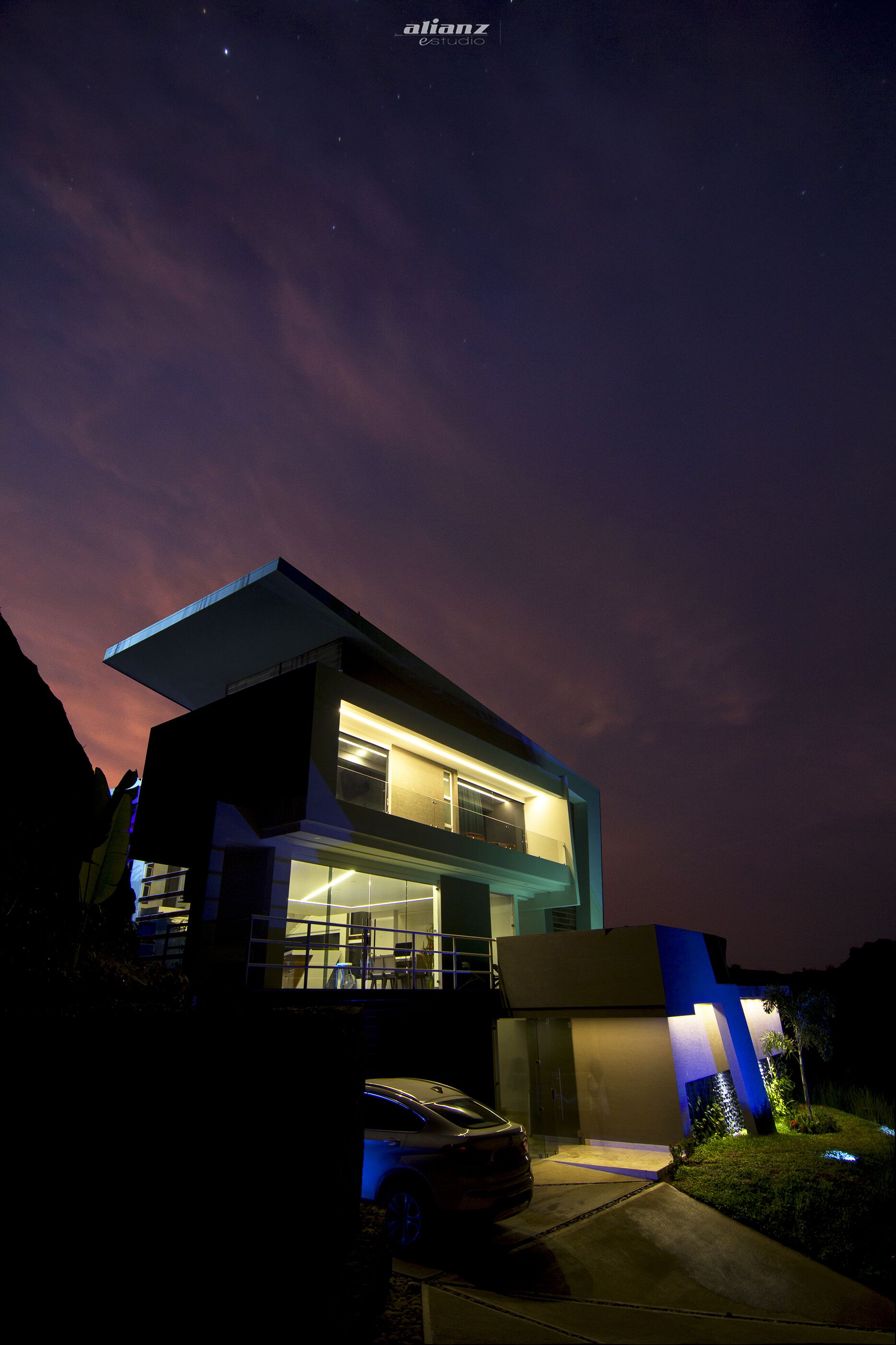 A modern two-story house with a flat roof and large windows, surrounded by a well-maintained lawn and a parked car.