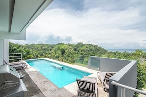 A rectangular swimming pool with clear blue water, surrounded by lounge chairs and a view of a lush green landscape and ocean.