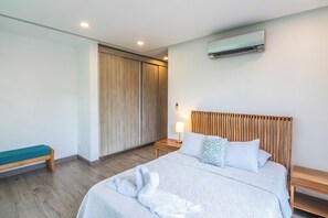 A hotel room with a bed, a bench, a bedside table, a nightstand, an air conditioning unit, and a wardrobe.