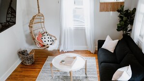 Ein Wohnzimmer mit einer schwarzen Couch, einem weißen runden Couchtisch, einem Hängekorbstuhl mit Kissen, einem gewebten Korb und einem gemusterten Teppich.