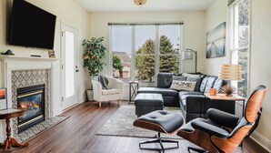 A living room with a fireplace, a television, a sofa, and a large window. - Wind Off The Lake - Updated Modern Townhouse (Manistee)