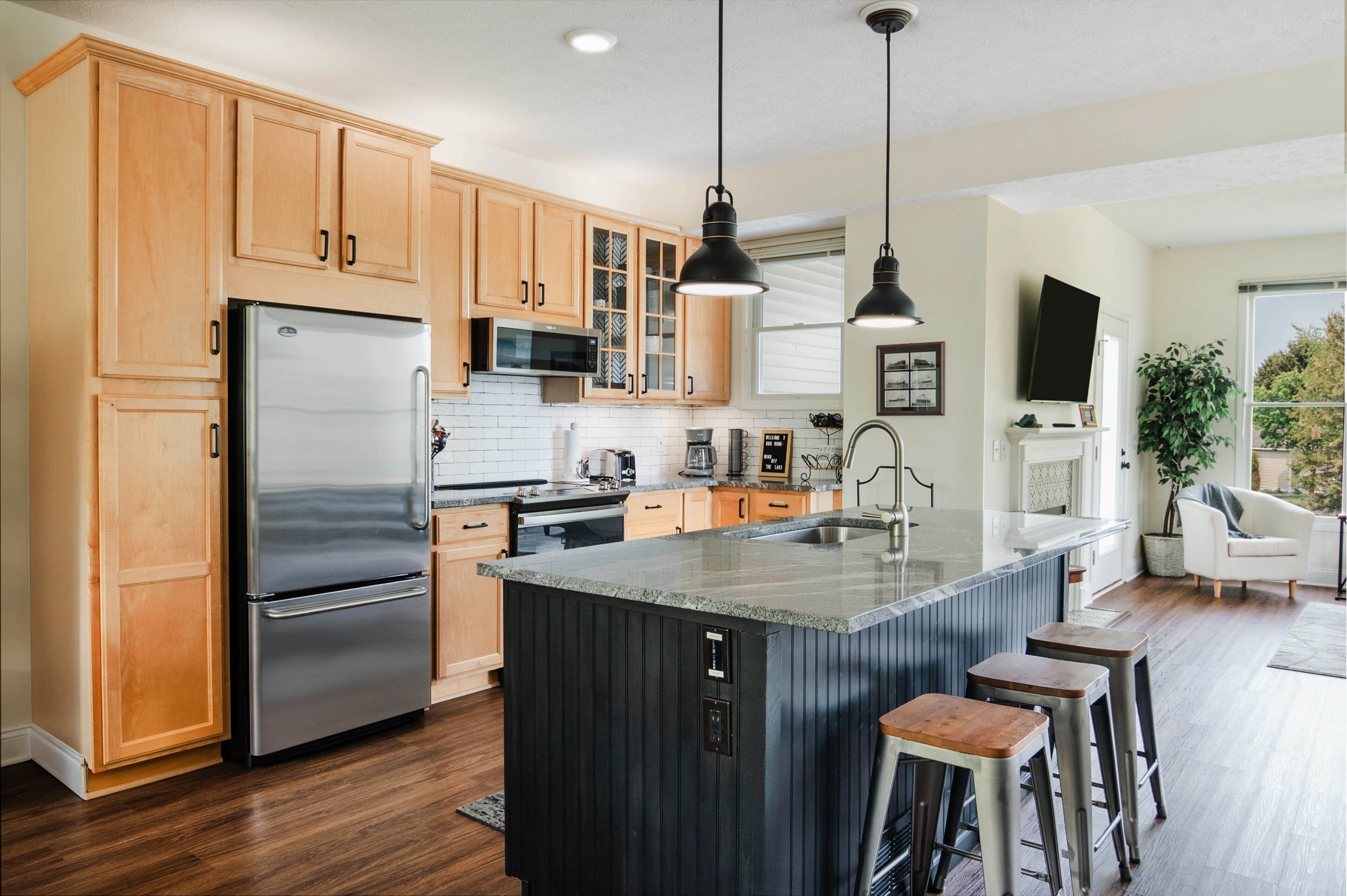 Een moderne keuken met houten kastjes, roestvrijstalen apparatuur, een centraal eiland met krukken, hanglampen en uitzicht op de omgeving.