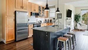 A modern kitchen with wooden cabinets, stainless steel appliances, a central island with stools, pendant lighting, and a view of the outdoors. - Wind Off The Lake - Updated Modern Townhouse (Manistee)