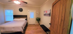 Une chambre à coucher comprenant un lit, un banc, une porte, une horloge, une fenêtre avec des stores, une plante et un tableau.