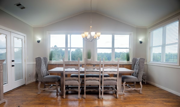 A dining room with a large table, four chairs, a chandelier, and large windows with a view of trees.