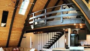 A loft-style living space with a high ceiling, exposed wooden beams, a staircase, a kitchen area, and a seating area with a glass table and chairs. - Modern A-Frame Cabin in Mammoth (Mammoth Lakes)