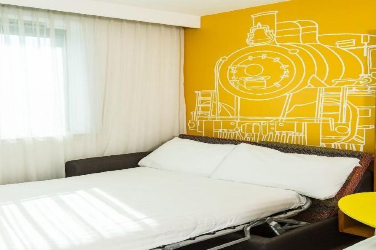 A hotel room with a bed, a yellow accent wall, and a window with curtains.