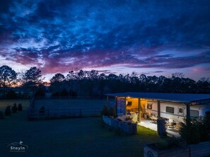 Una caravana bien iluminada con un espacio exterior cubierto, rodeada de árboles y un campo cercado.