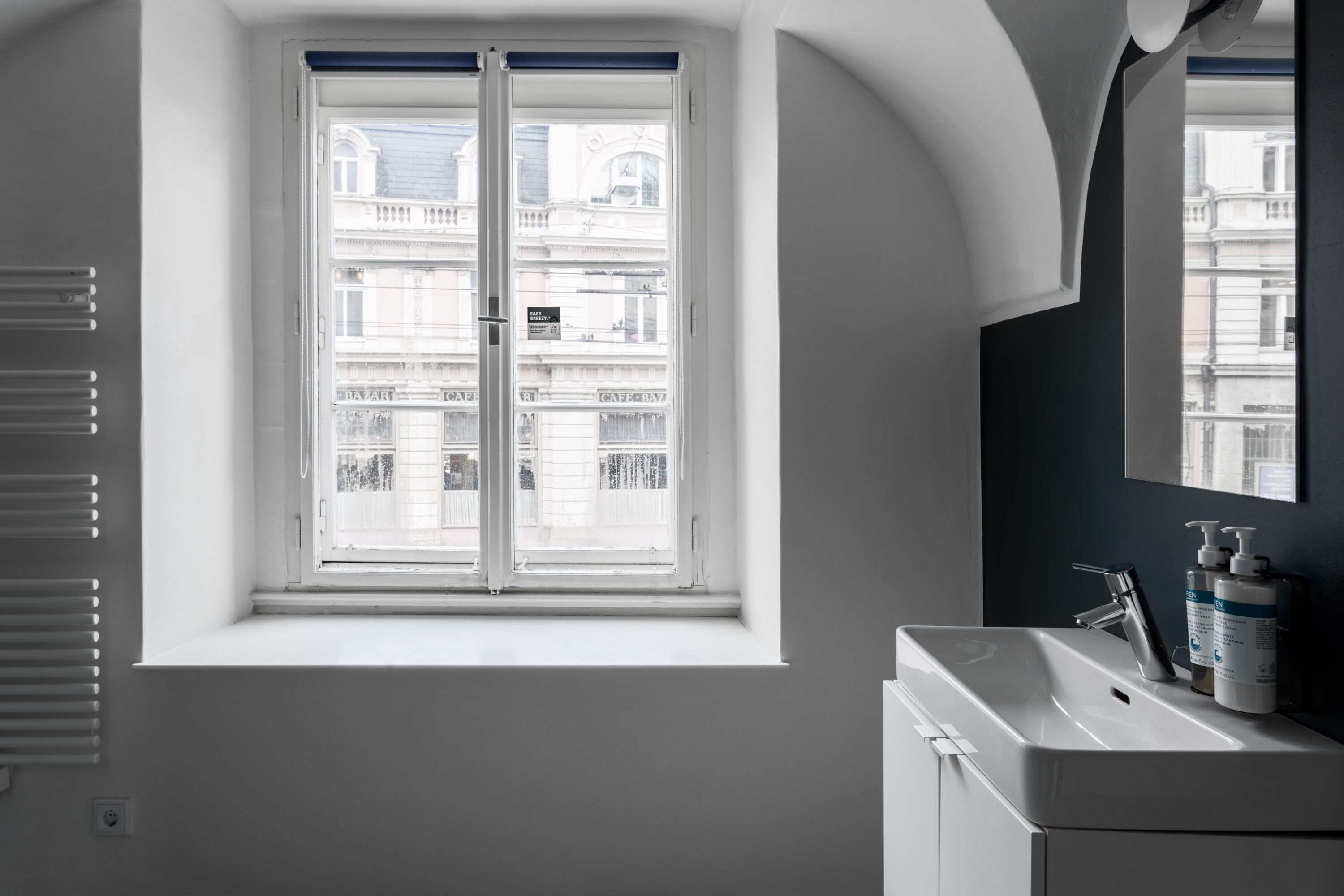 Une salle de bain avec un lavabo blanc, un miroir et une fenêtre donnant sur des immeubles.