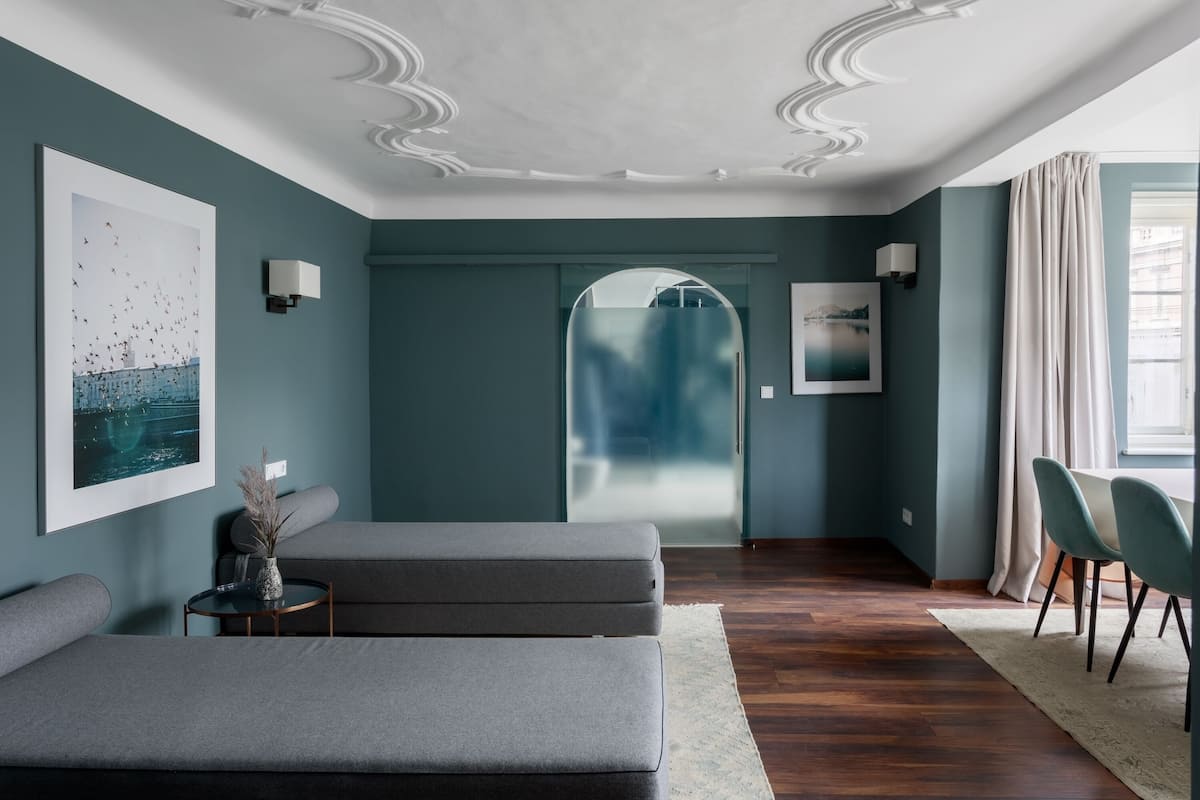 A modern room with teal walls, a wooden floor, and a large window with curtains. There are two grey sofas, a small round table with a vase, and a wall-mounted lamp.