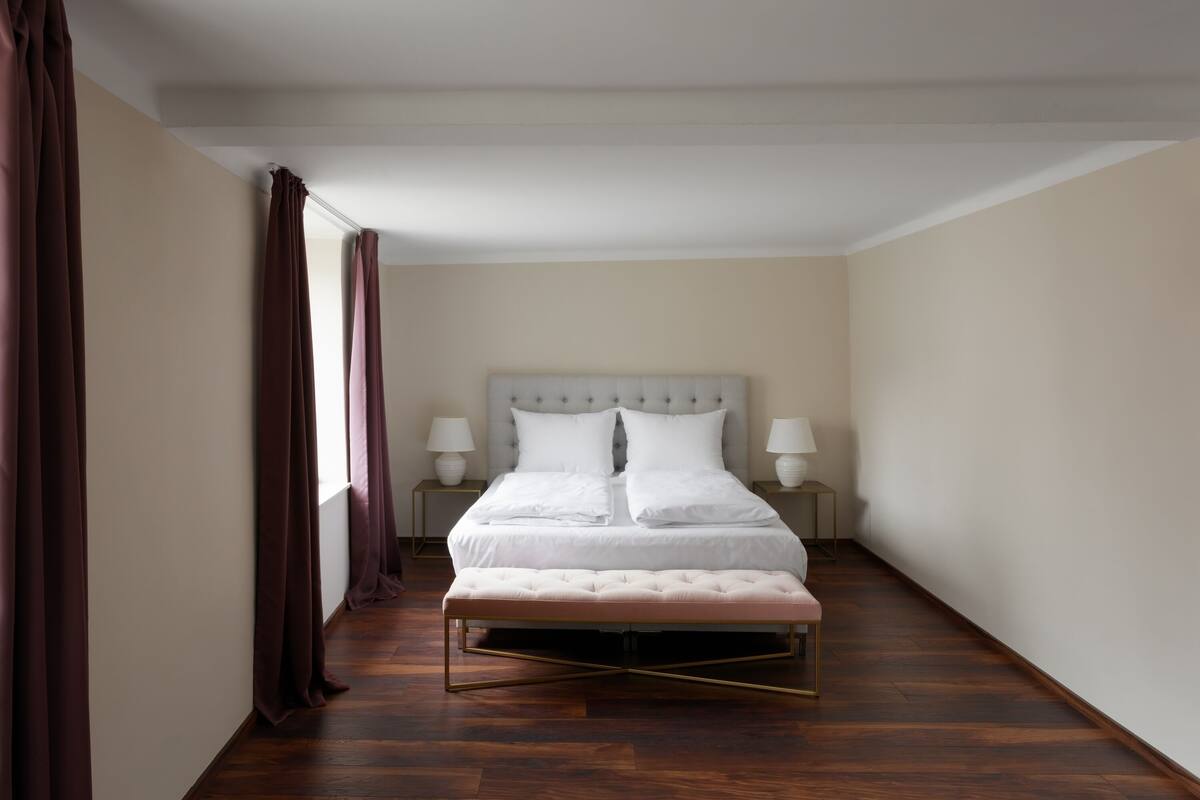A bedroom with a large bed, two bedside tables with lamps, a bench, and a view of the outdoors through curtains.