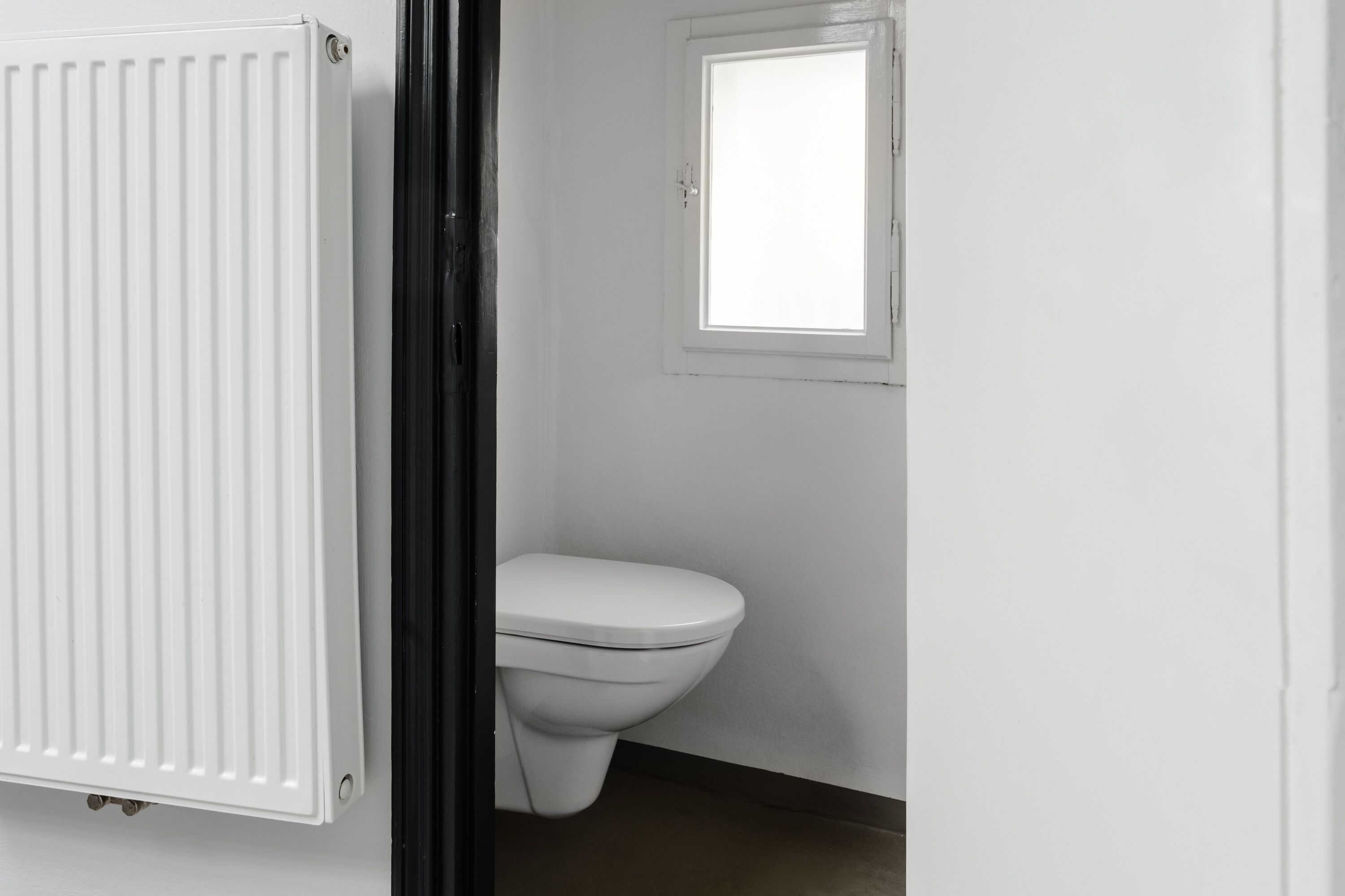 Un inodoro blanco con una ventana arriba, un radiador blanco y un suelo oscuro.