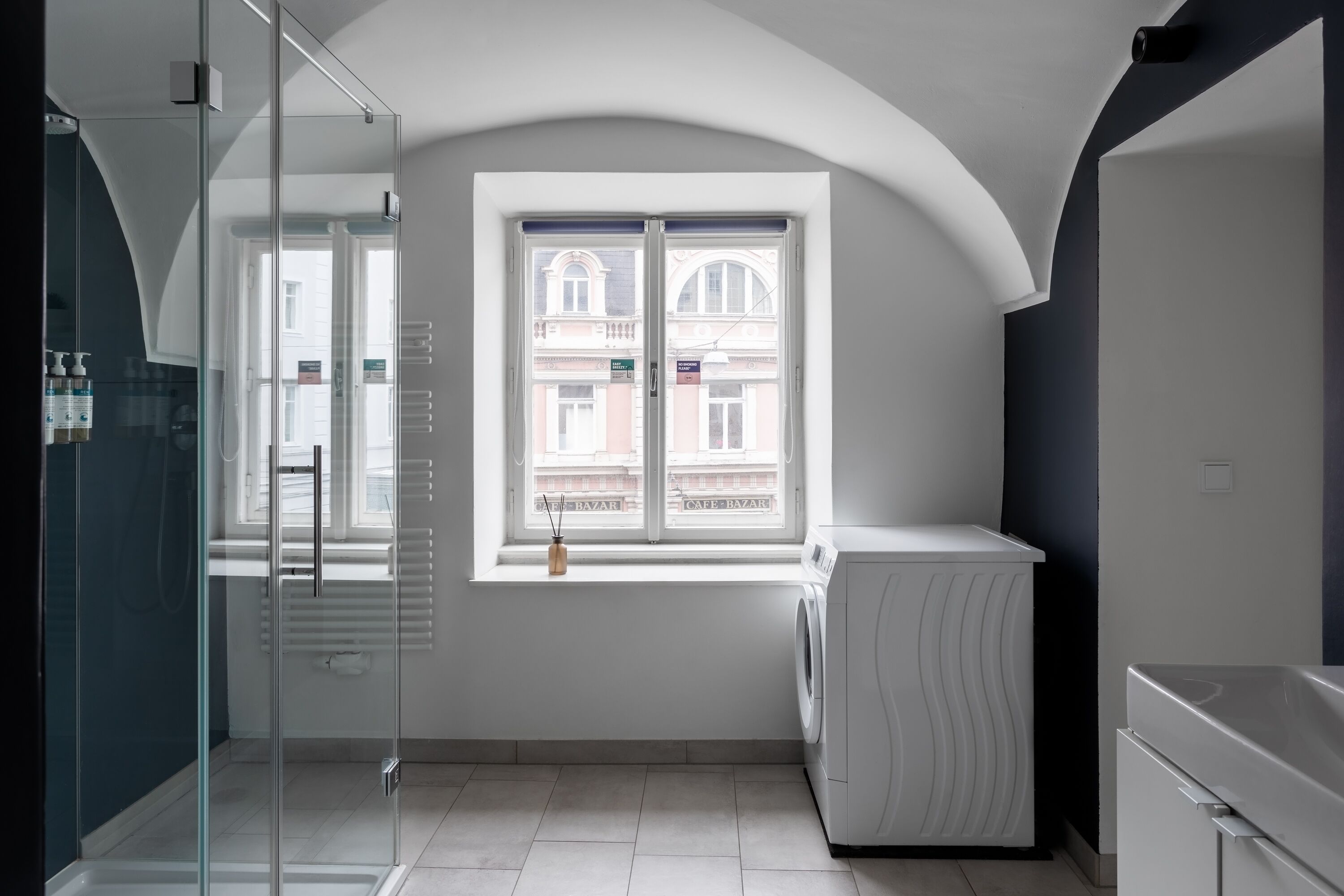 Une salle de bain moderne et compacte, dotée d’une douche à parois de verre, d’un lave-linge encastré et d’une fenêtre donnant sur les bâtiments environnants.