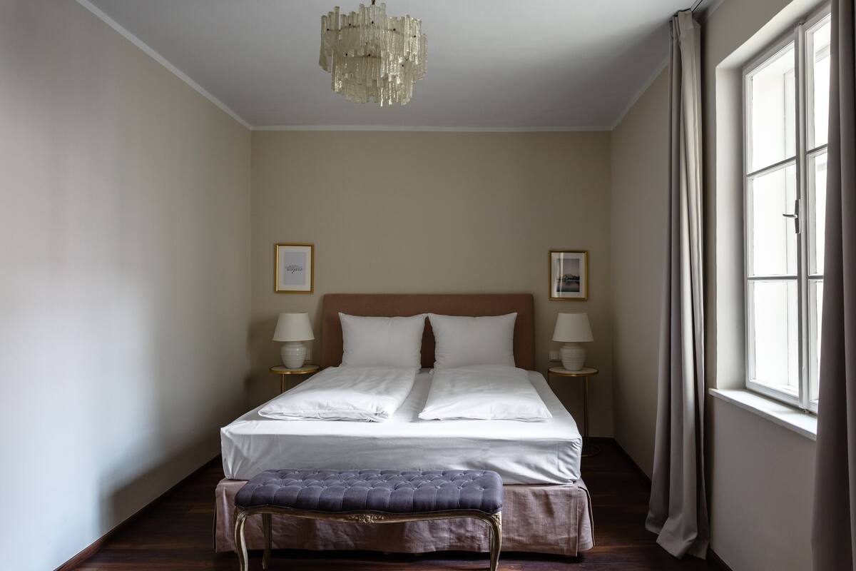 A neatly arranged bedroom with a large bed, bedside lamps, a chandelier, and a window with curtains.
