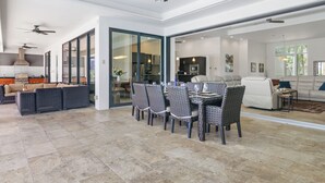 Amplia sala de estar con una mesa de comedor, sillas y un sofá. Hay ventiladores de techo, una cocina al fondo y un gran ventanal corredero que da a otra habitación.