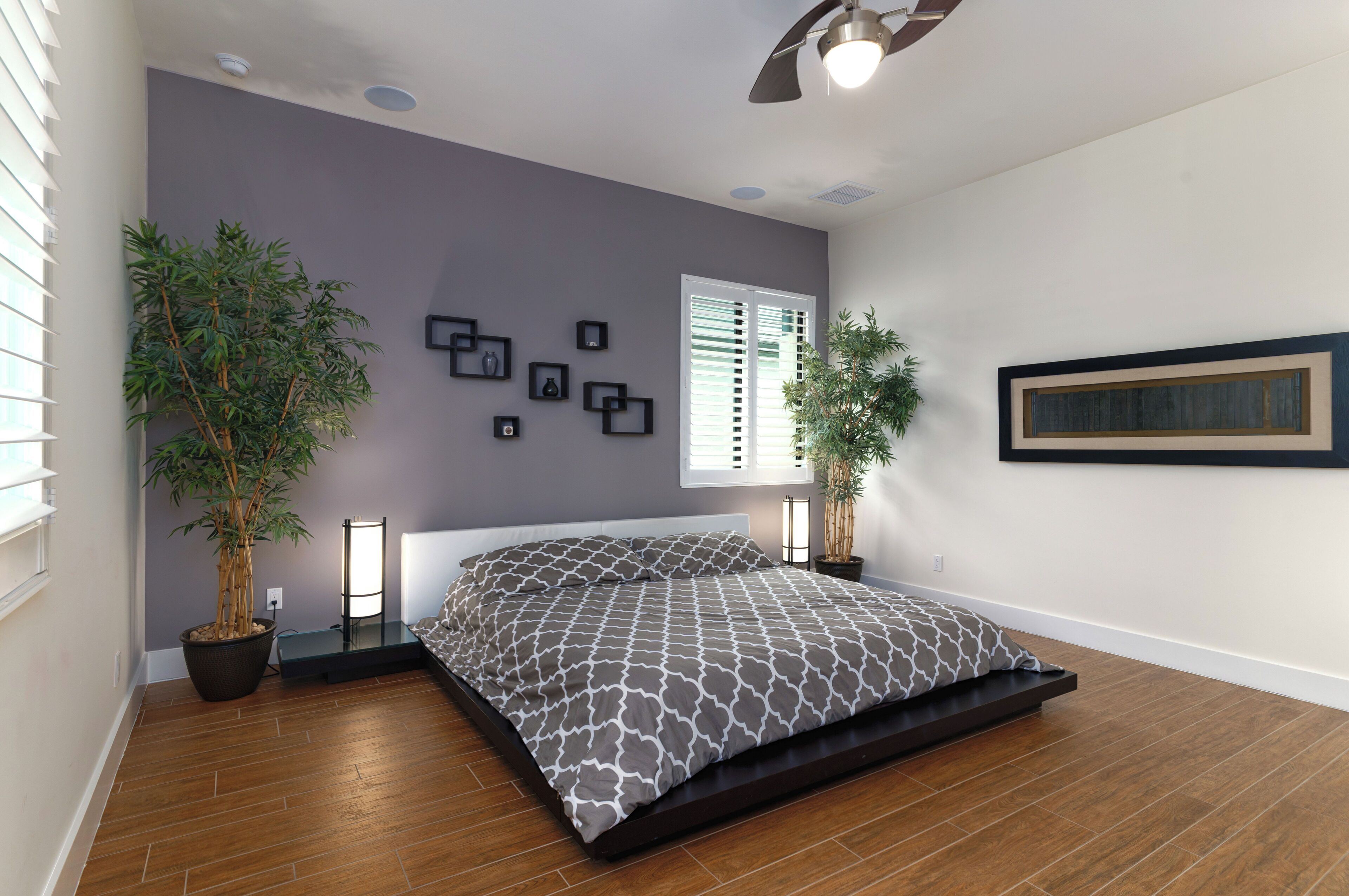 A bedroom with a bed, two potted plants, a ceiling fan, and a window with blinds.