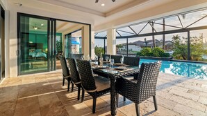Un comedor con una mesa y sillas de vidrio, un ventilador de techo y vista a una piscina.