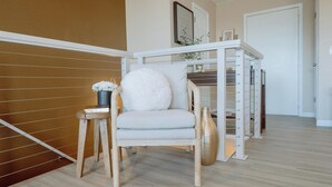 Una habitación moderna con suelo de madera, una silla blanca con un cojín mullido, una mesita de noche con un jarrón y un estante blanco adosado a la pared.
